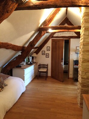 A cozy attic bedroom with exposed wooden beams, a stone wall, a wooden door, a bed, a chair, and a desk with a lamp.