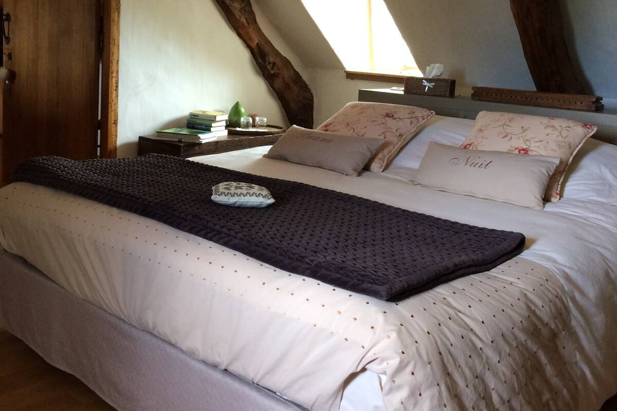 Un dormitorio con una cama grande, vigas de madera, tragaluces y una mesita de noche con libros y una lámpara.