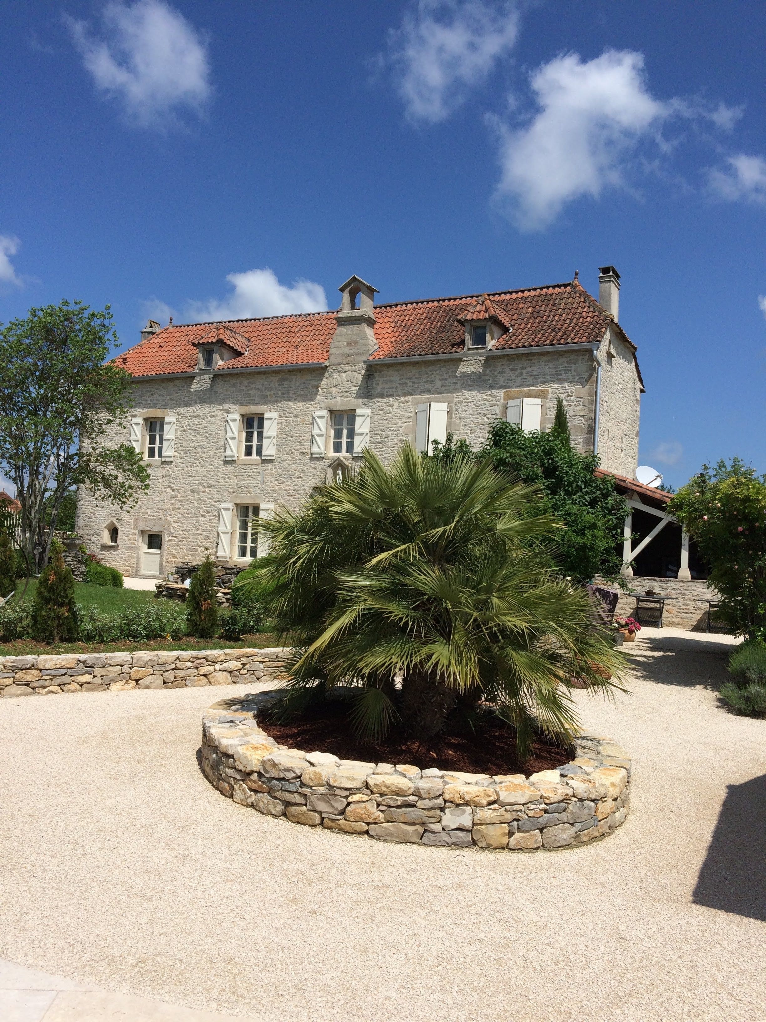 Un bâtiment en pierre surmonté d’un toit de tuiles rouges, un parterre circulaire en pierre et un ciel d’un bleu limpide.