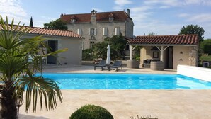 Una piscina con sillas de descanso y un área de estar techada, rodeada de edificios de piedra y vegetación.