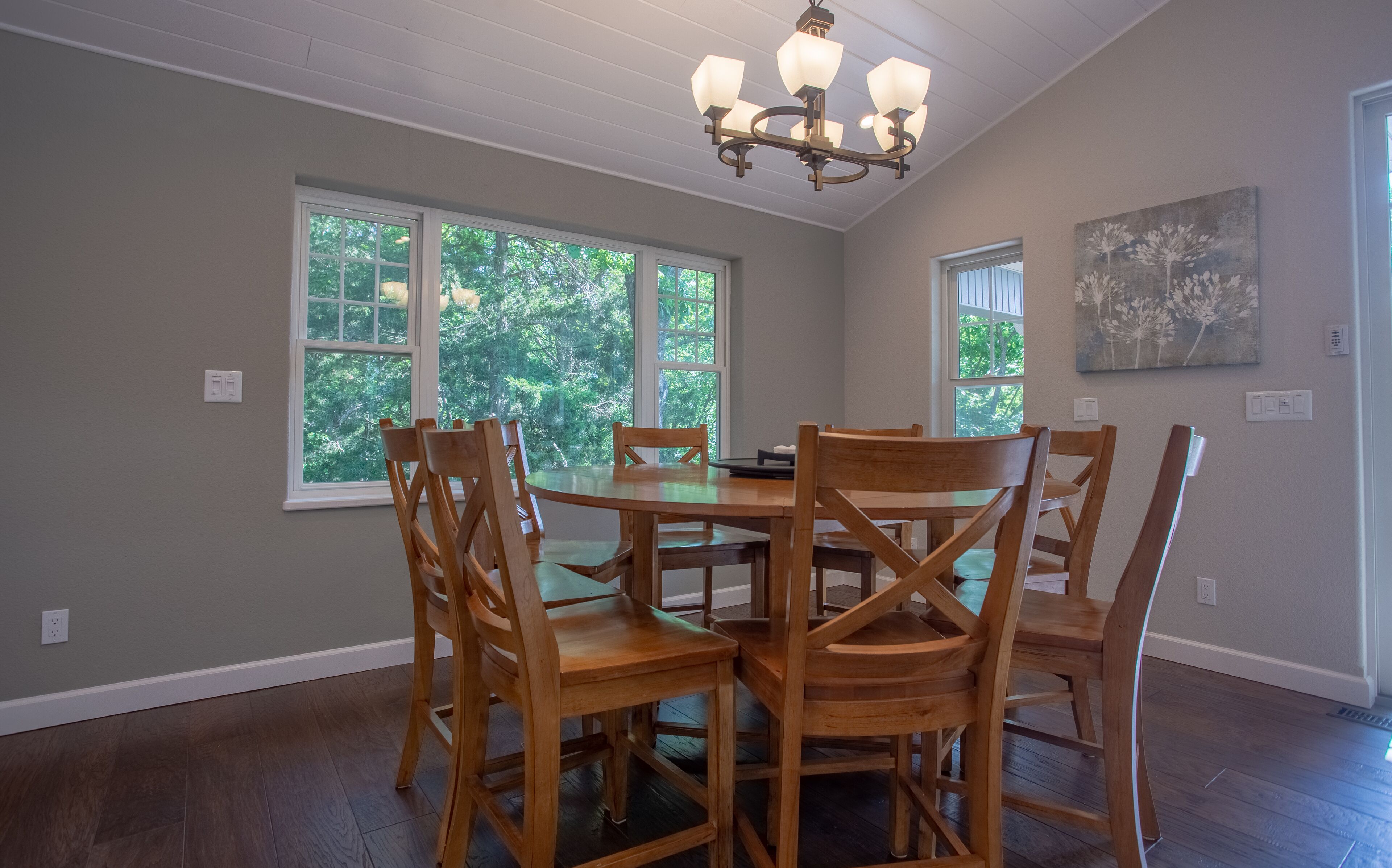 A dining area with a round table and wooden chairs, large windows with a view of trees, and a chandelier.