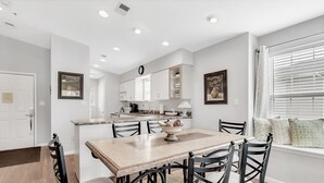 A modern kitchen with white cabinets, a wooden dining table, and chairs. There is a window with blinds and a door leading to another room. - Gorgeous Greens 2 Bedroom Condo by RedAwning (Branson)