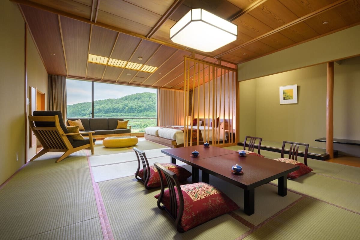 Ein traditioneller japanischer Raum mit Tatami-Böden, einem niedrigen Tisch mit Teesets, einer Couch und Blick auf einen bewaldeten Hügel.