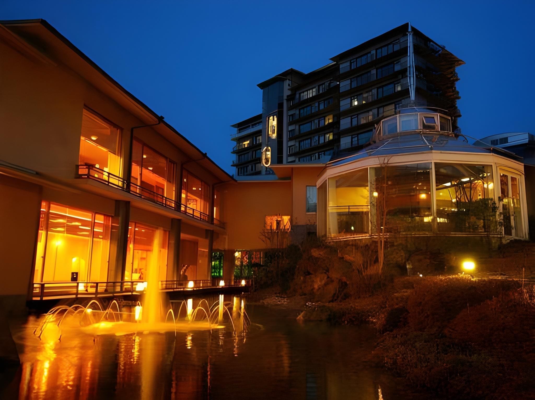Ein Hotel mit modernem Design, das durch eine große Glasarchitektur und eine sich spiegelnde Wasserspiele im Vordergrund gekennzeichnet ist.