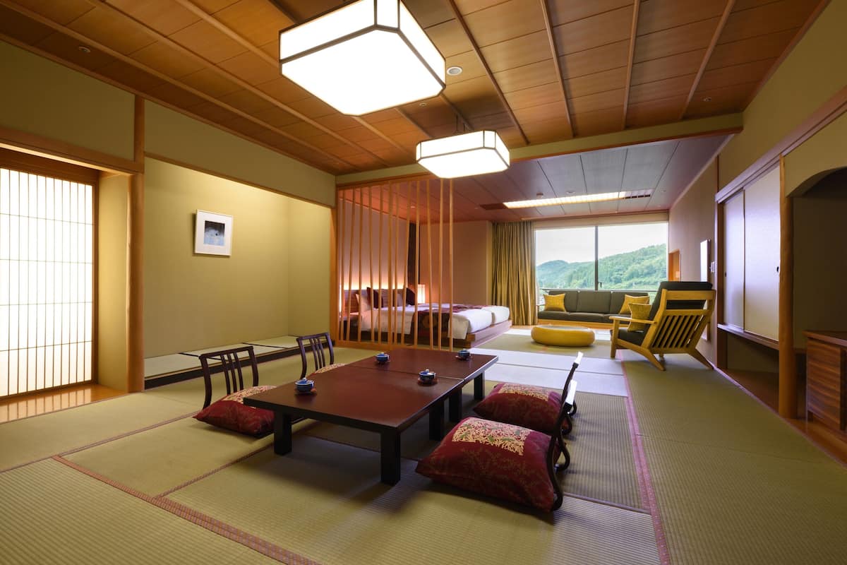 Ein traditioneller japanischer Raum mit Tatami-Bodenbelag, einem niedrigen Tisch mit Kissen, einer Couch und Blick auf eine gebirgige Landschaft.