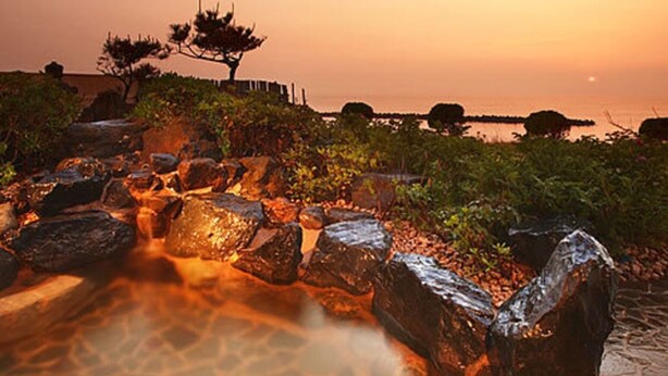 Una fuente de piedra con rocas y una pequeña cascada, rodeada de vegetación y una vista distante del mar al atardecer.
