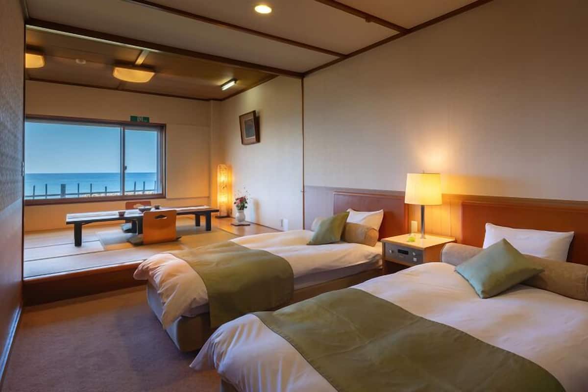 Habitación de hotel con dos camas, una ventana con vistas, una mesita de noche con lámpara y un banco junto a la ventana.