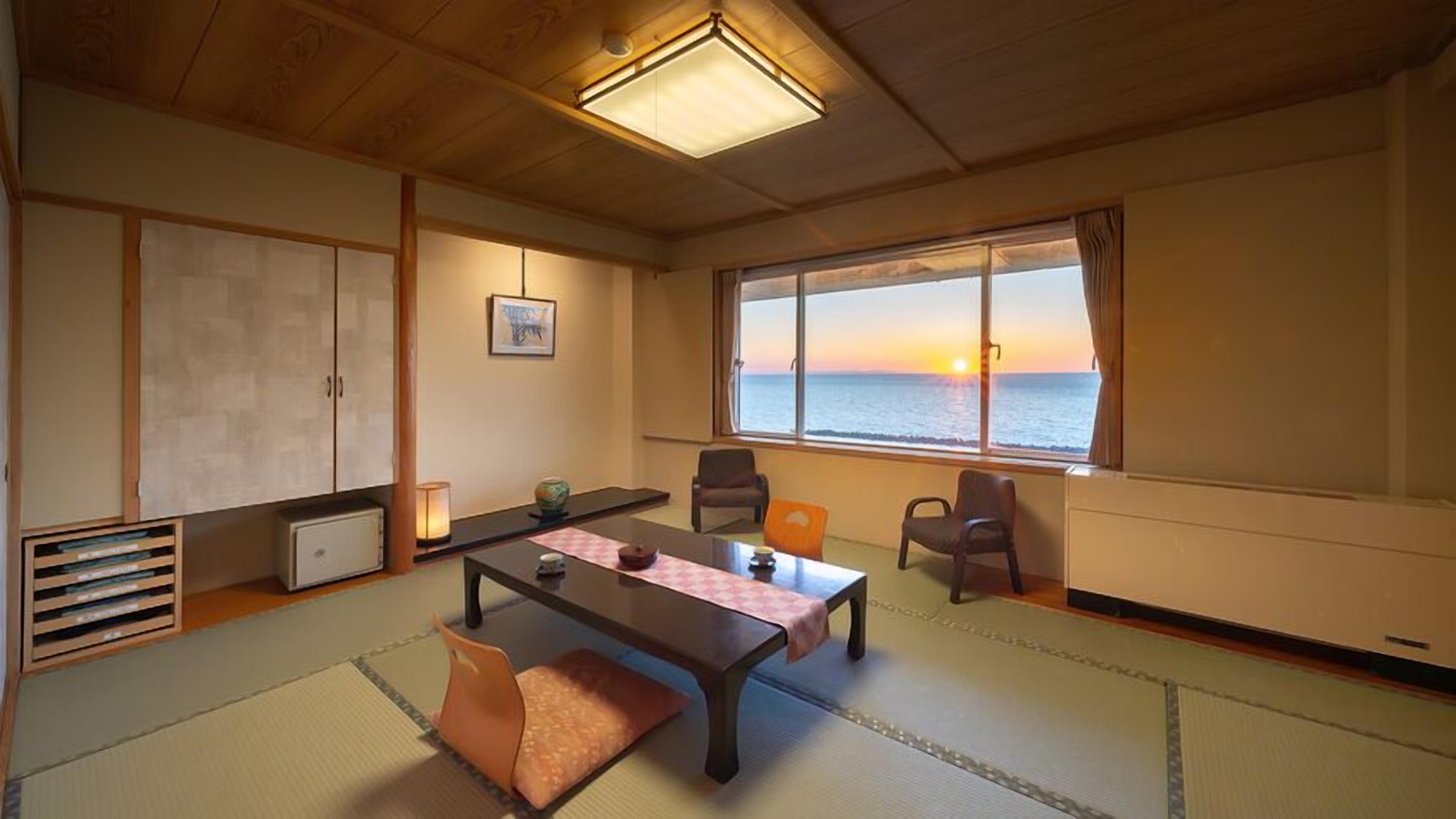 Una habitación japonesa tradicional con suelo de tatami, una mesa baja, sillas, vista al mar al atardecer y una lámpara en el techo.