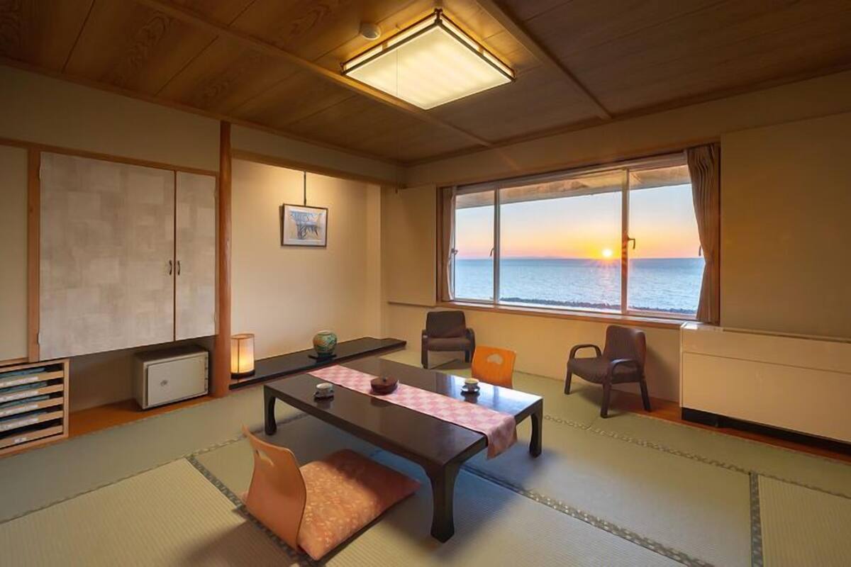Una habitación japonesa tradicional con suelo de tatami, una mesa baja, sillas, vista al mar al atardecer y una lámpara en el techo.