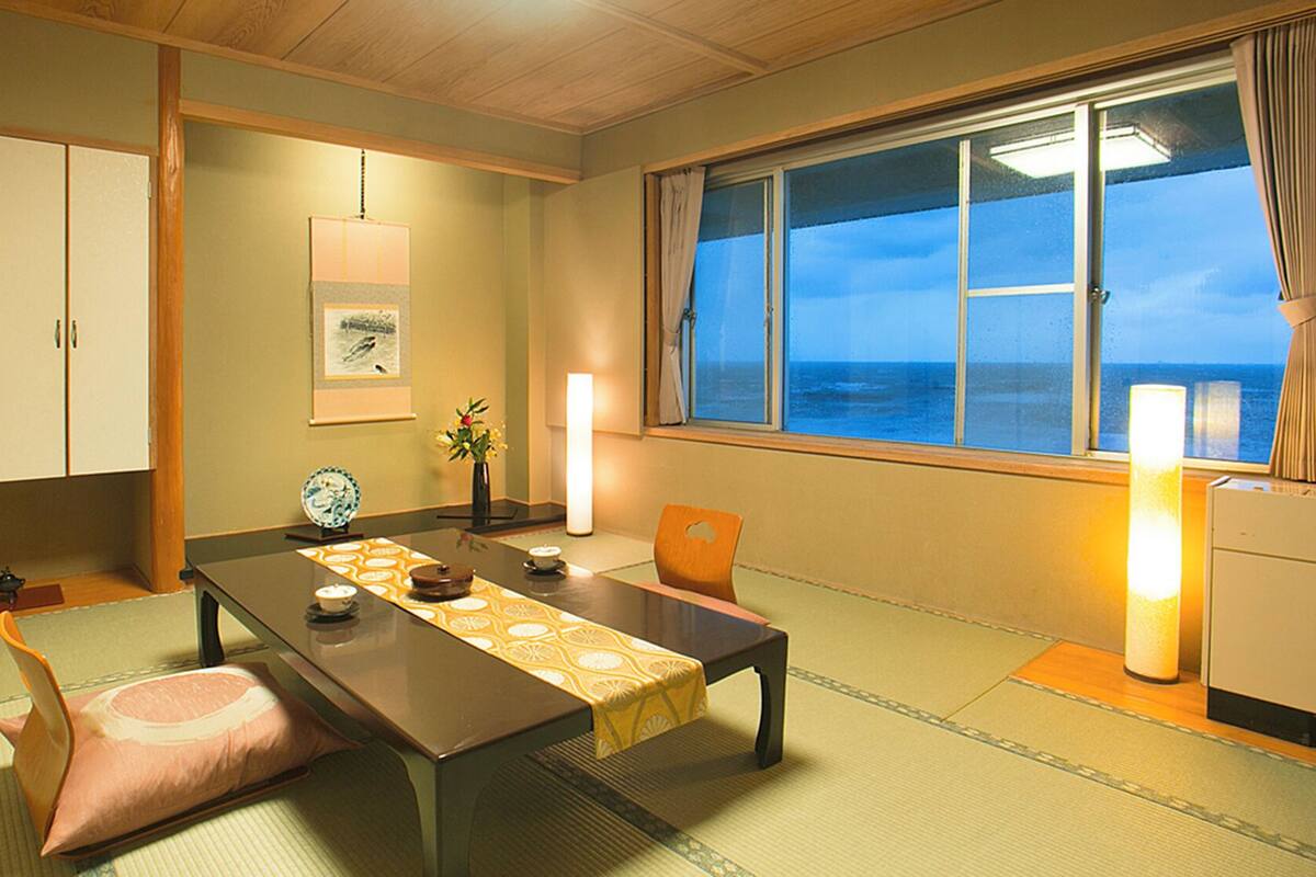 Una habitación japonesa tradicional con suelo de tatami, una mesa baja, vistas al mar y un ventanal grande.