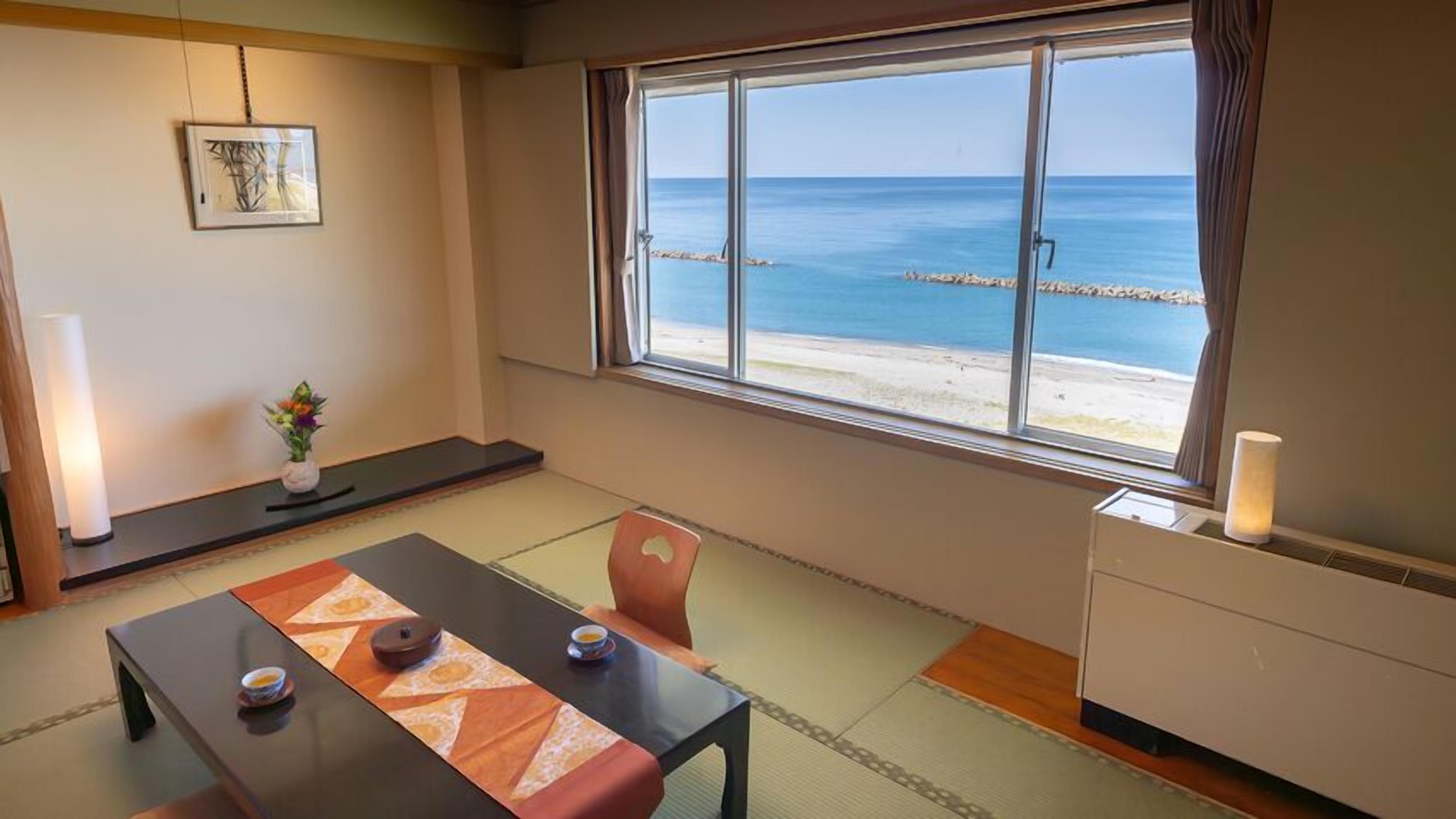 Una habitación con suelo de tatami, una mesa baja con cojín, una silla, una pequeña mesa con una vela y un ventanal con vistas al mar y un muelle.