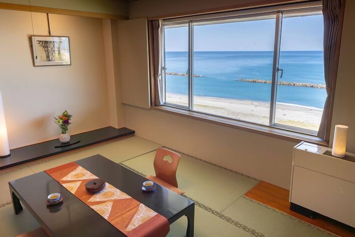 Una habitación con suelo de tatami, una mesa baja con cojín, una silla, una pequeña mesa con una vela y un ventanal con vistas al mar y un muelle.