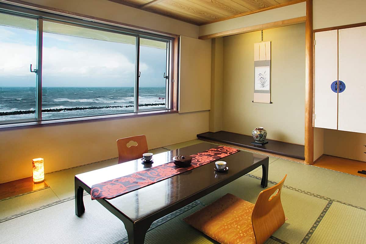 Una habitación japonesa tradicional con suelo de tatami, una mesa baja con un tapete rojo y vistas al océano, además de un puertas correderas.