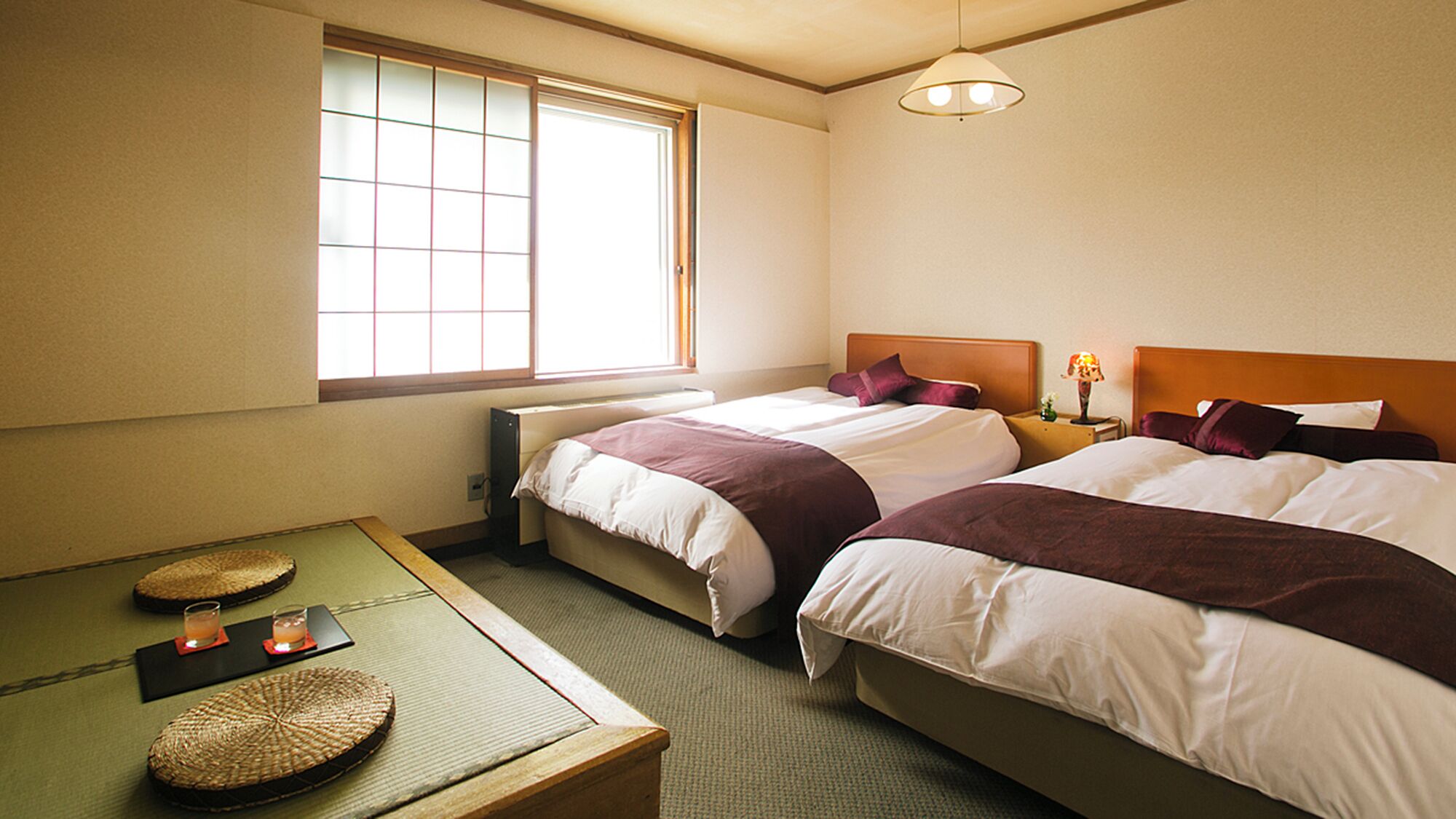 Habitación de hotel con dos camas, una mesita con un tapete, una lámpara y una ventana con persianas.