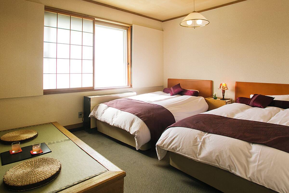 Habitación de hotel con dos camas, una mesita con un tapete, una lámpara y una ventana con persianas.
