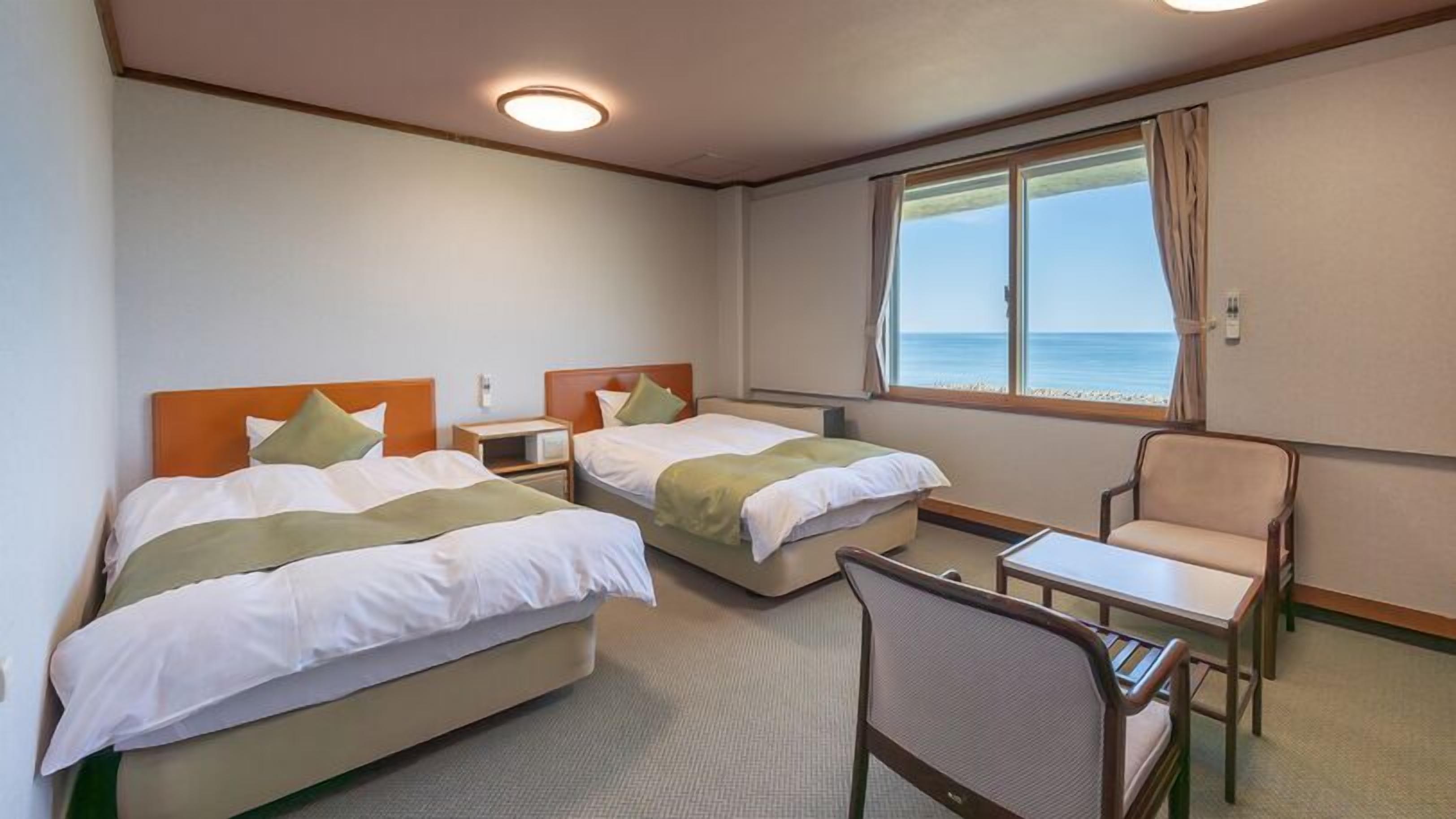 Habitación de hotel con dos camas, una mesita, una silla y ventana con vista al mar.