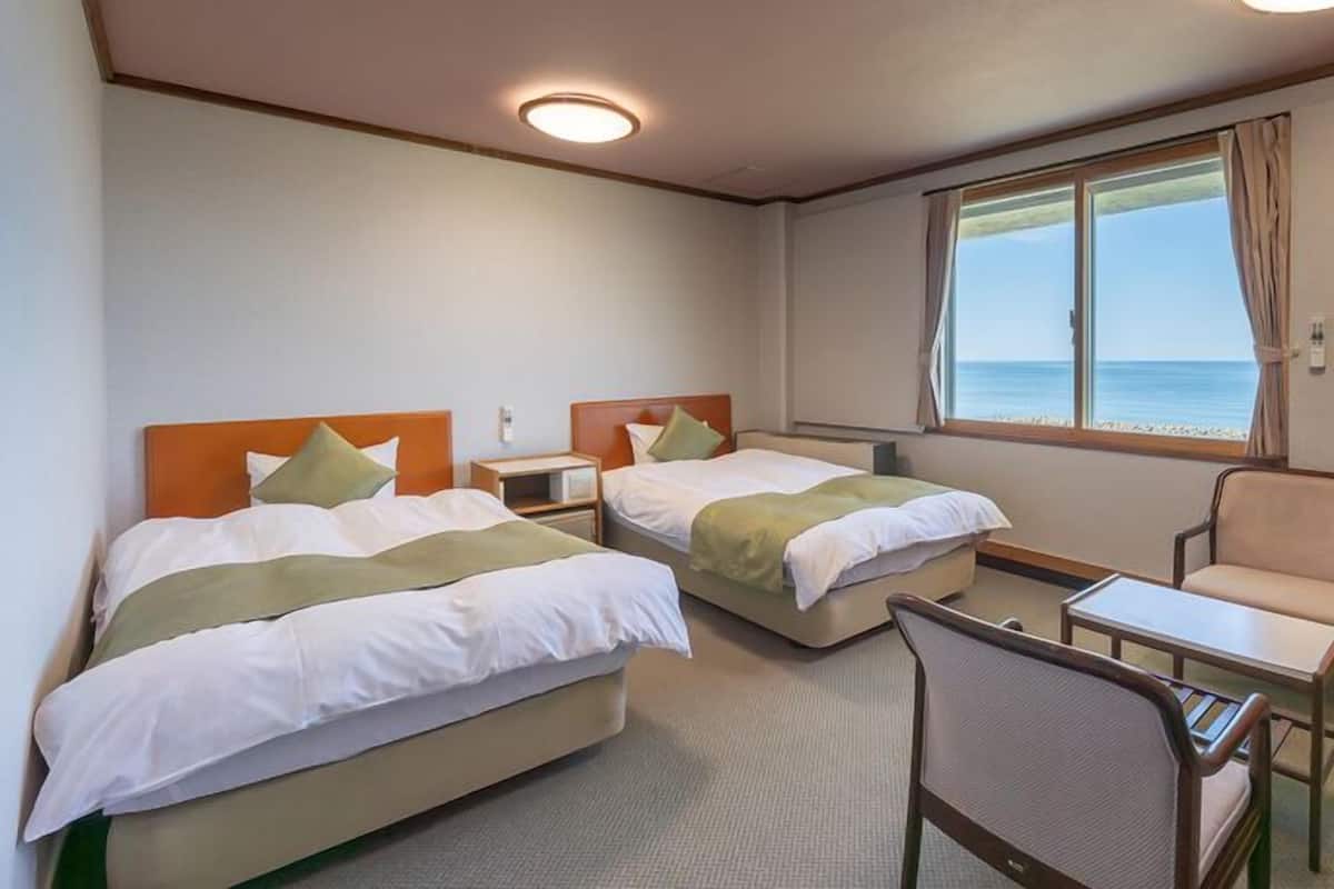 Habitación de hotel con dos camas, una mesita, una silla y ventana con vista al mar.