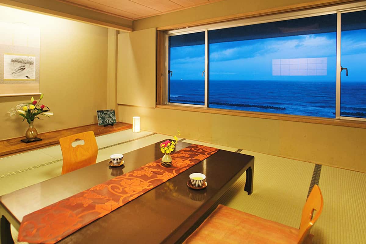 Una habitación con una mesa de madera, sillas, un jarrón con flores, un cuadro en la pared y vistas al mar a través de una ventana.
