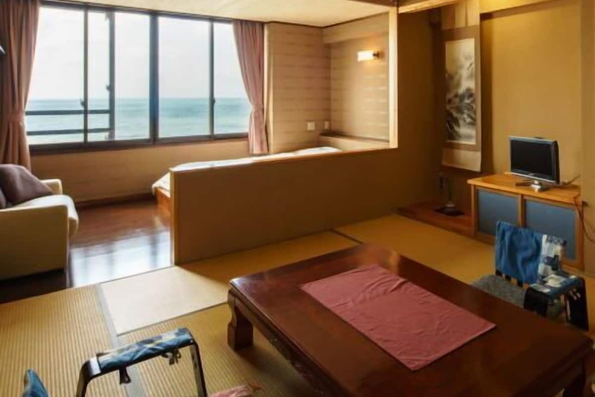 Una habitación con un ventanal grande, un sofá, una mesa de madera, un televisor y un pequeño rincón de descanso.