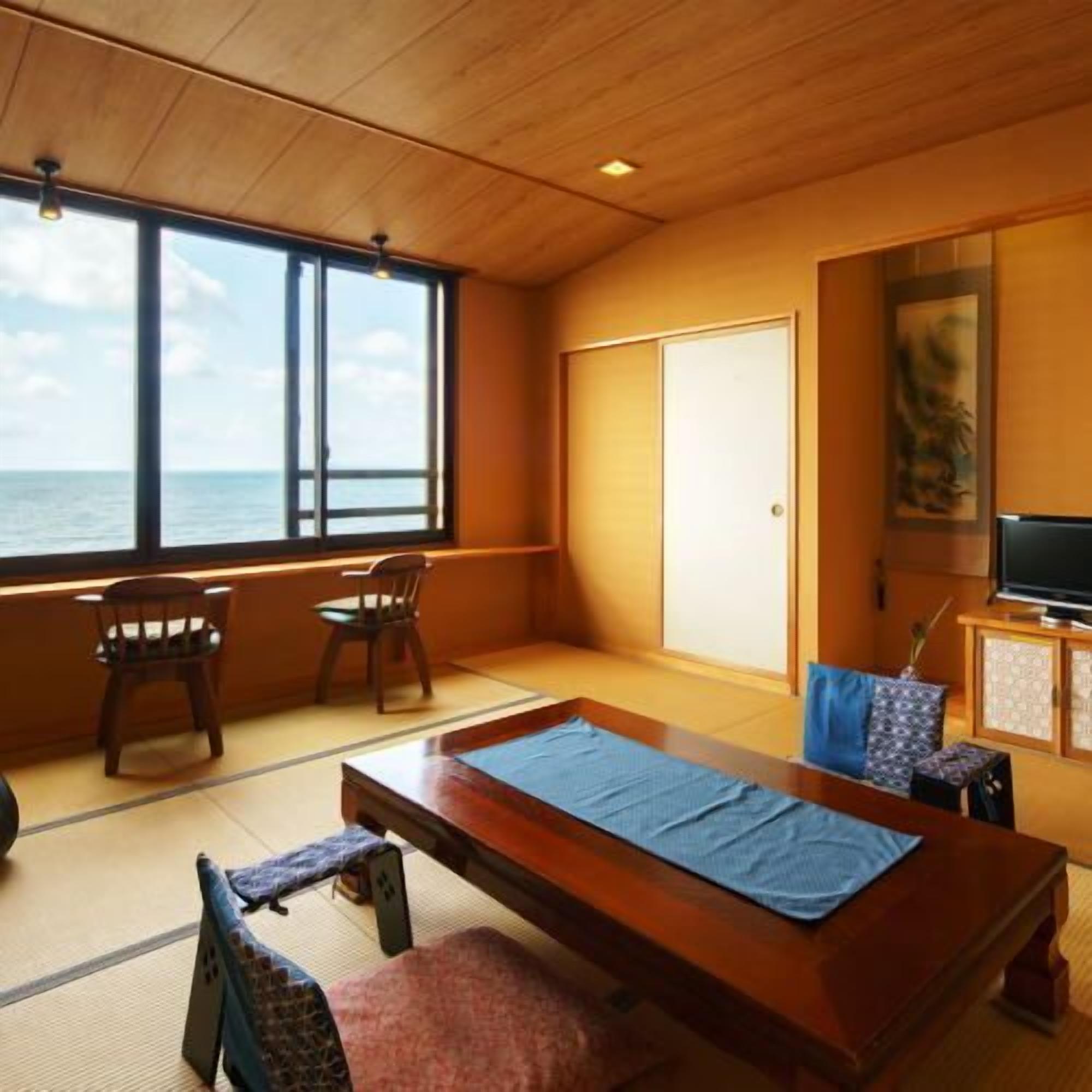 A room with tatami flooring, a wooden table, chairs, a television, and a view of the sea.