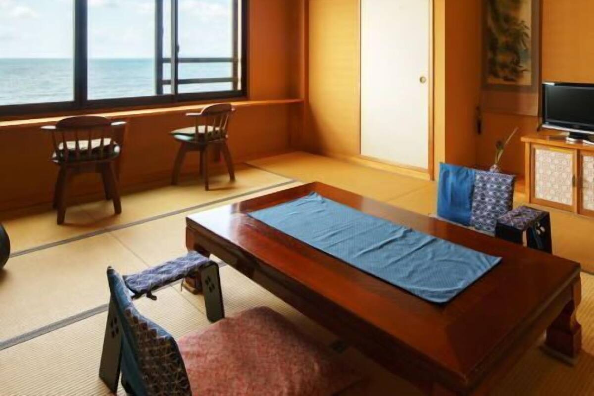 Una habitación con suelo de tatami, una mesa de madera, sillas, televisión y vista al mar.