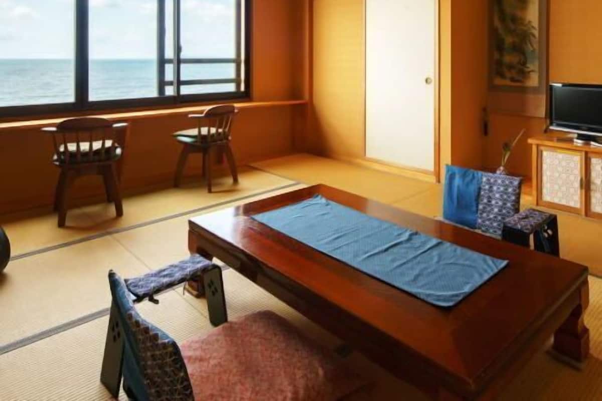 Una habitación con suelo de tatami, una mesa de madera, sillas, televisión y vista al mar.