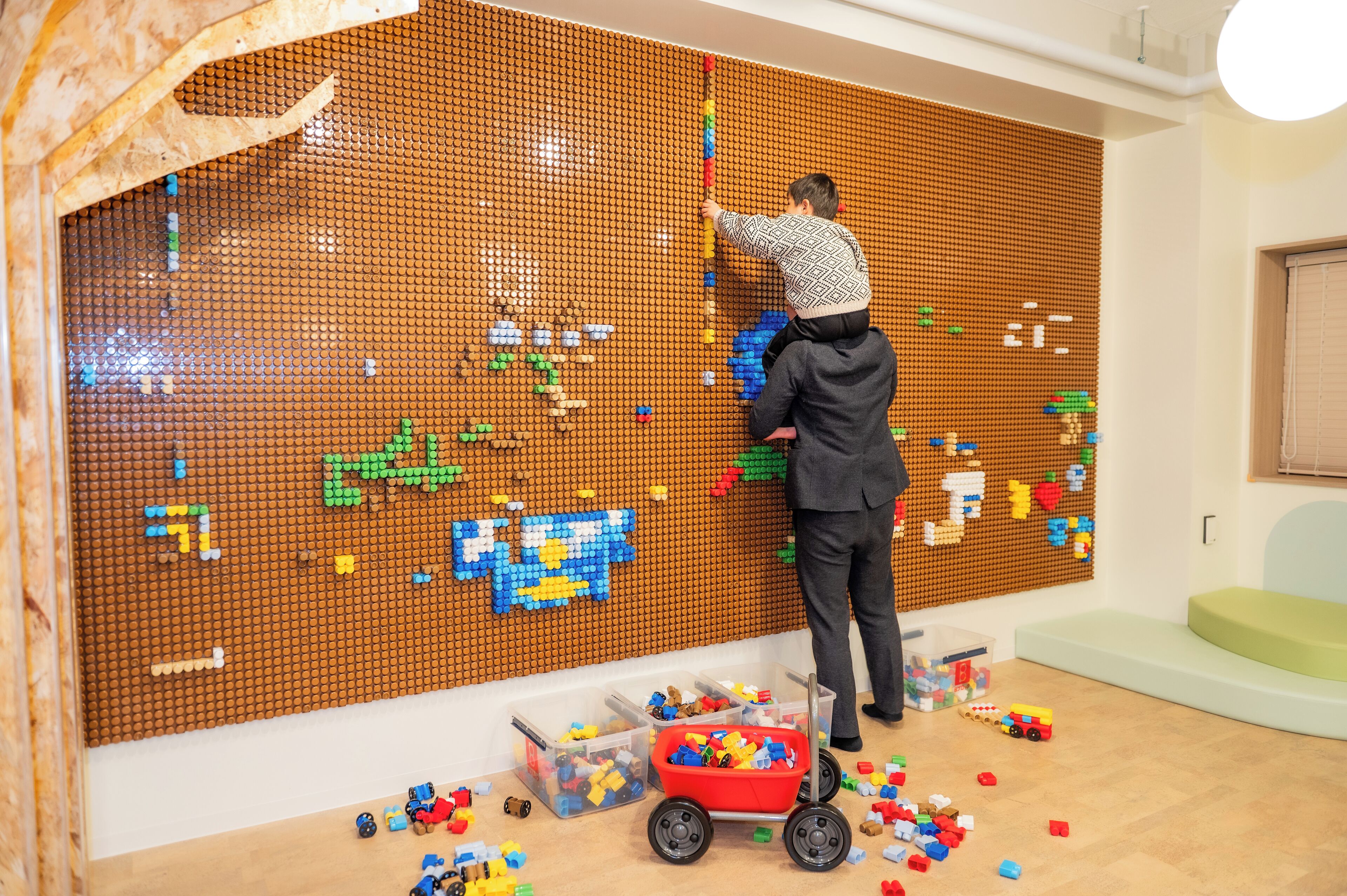 Une pièce avec un grand mur décoré de briques LEGO, un petit chariot rouge rempli de jouets et des briques LEGO éparpillées sur le sol.