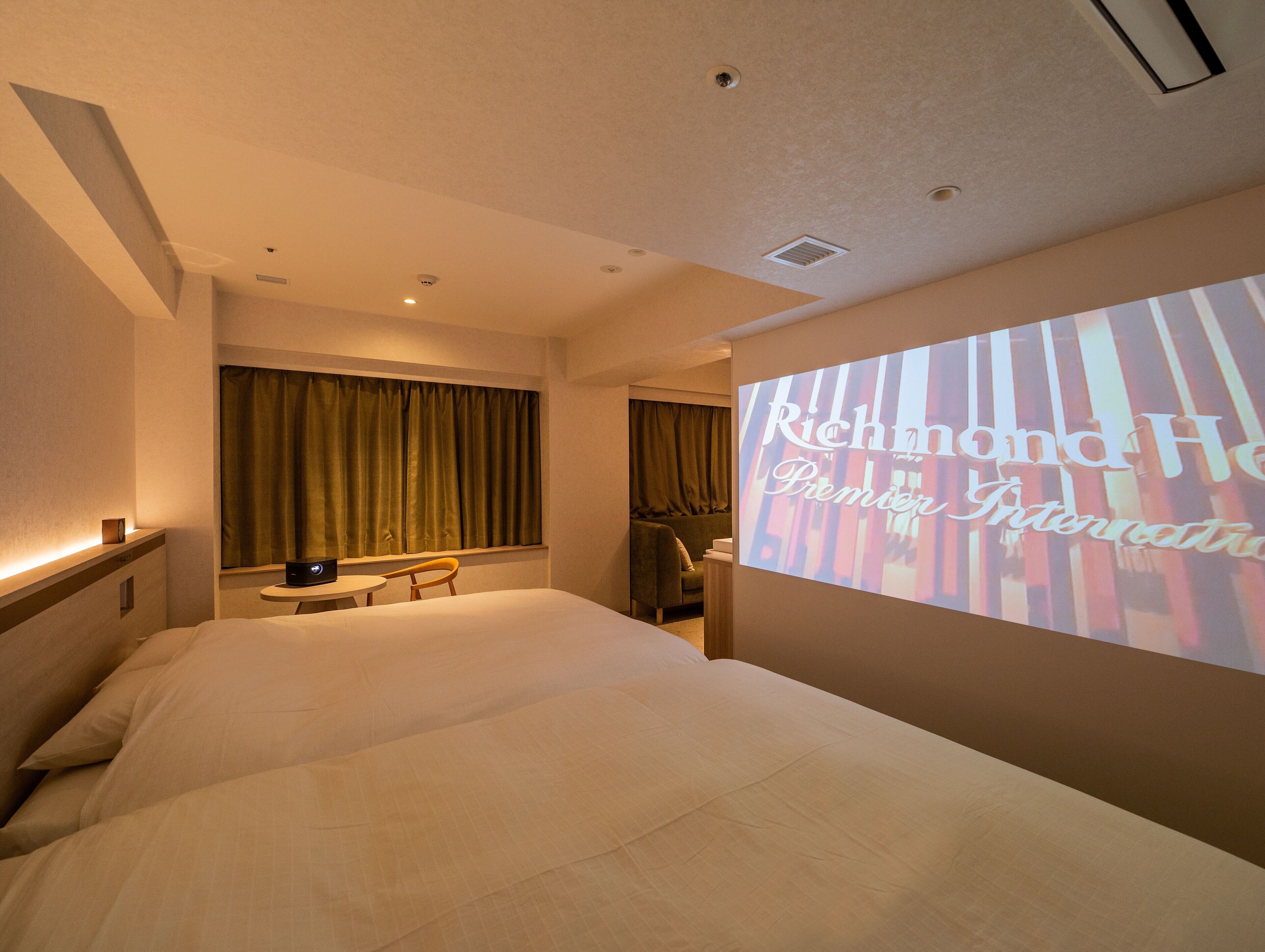 A hotel room with a bed, a desk, and a projection screen displaying "Richmond Hotel Premier International".