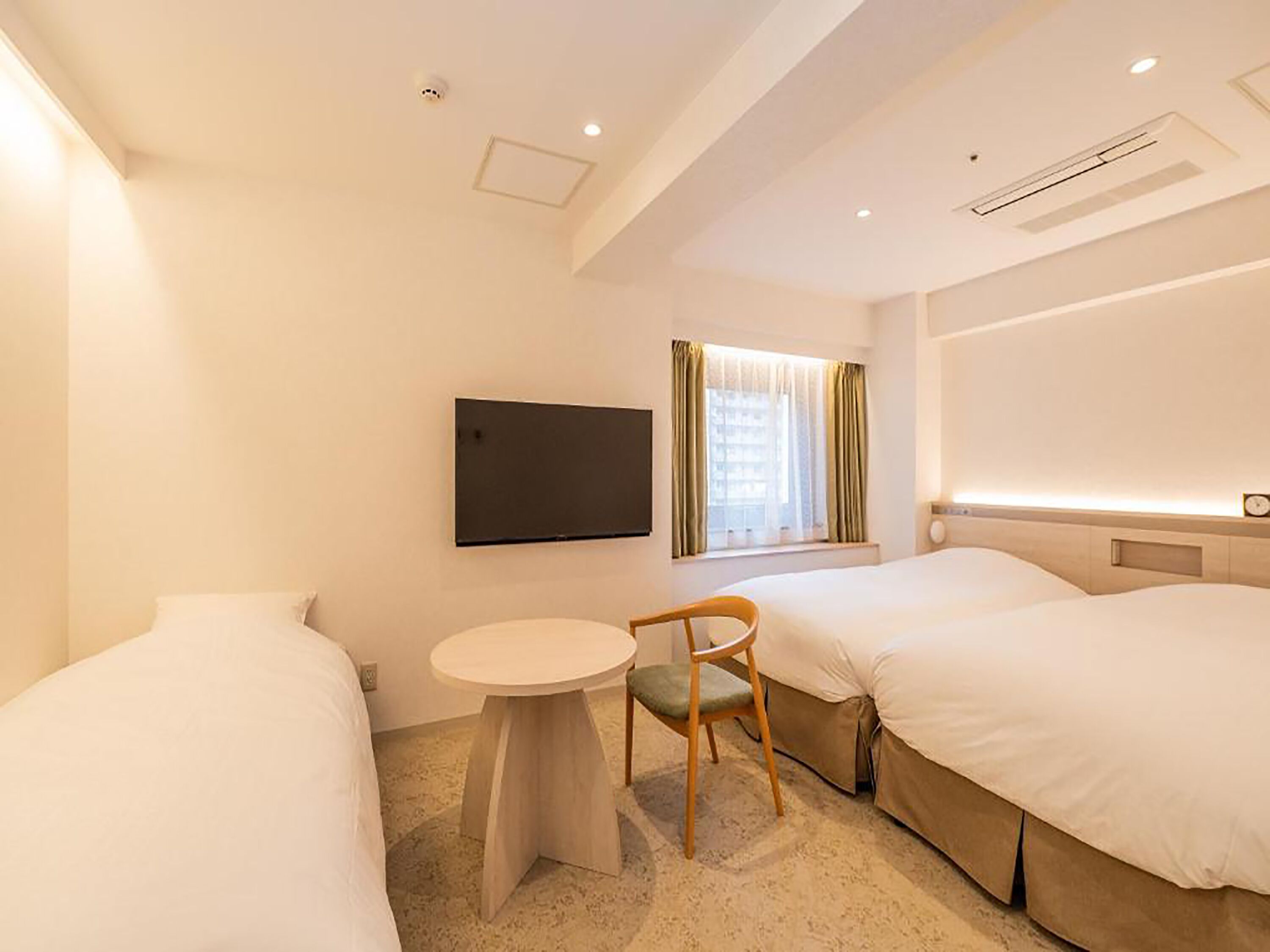 Quarto de hotel com duas camas, uma mesa redonda pequena, uma cadeira, uma televisão na parede e uma janela com cortinas.