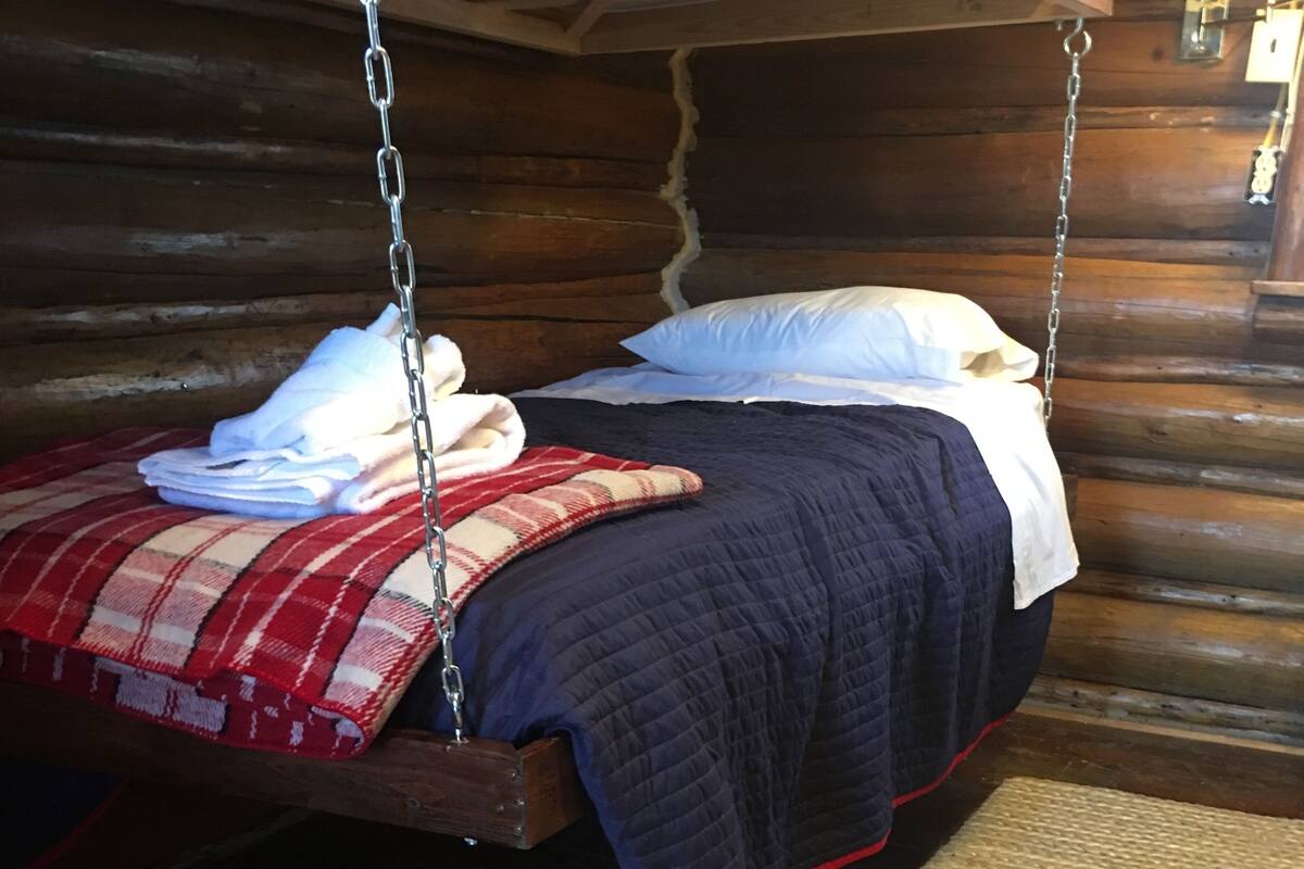 A rustic log cabin room with a bed suspended from the ceiling, a woven rug, and wooden walls.