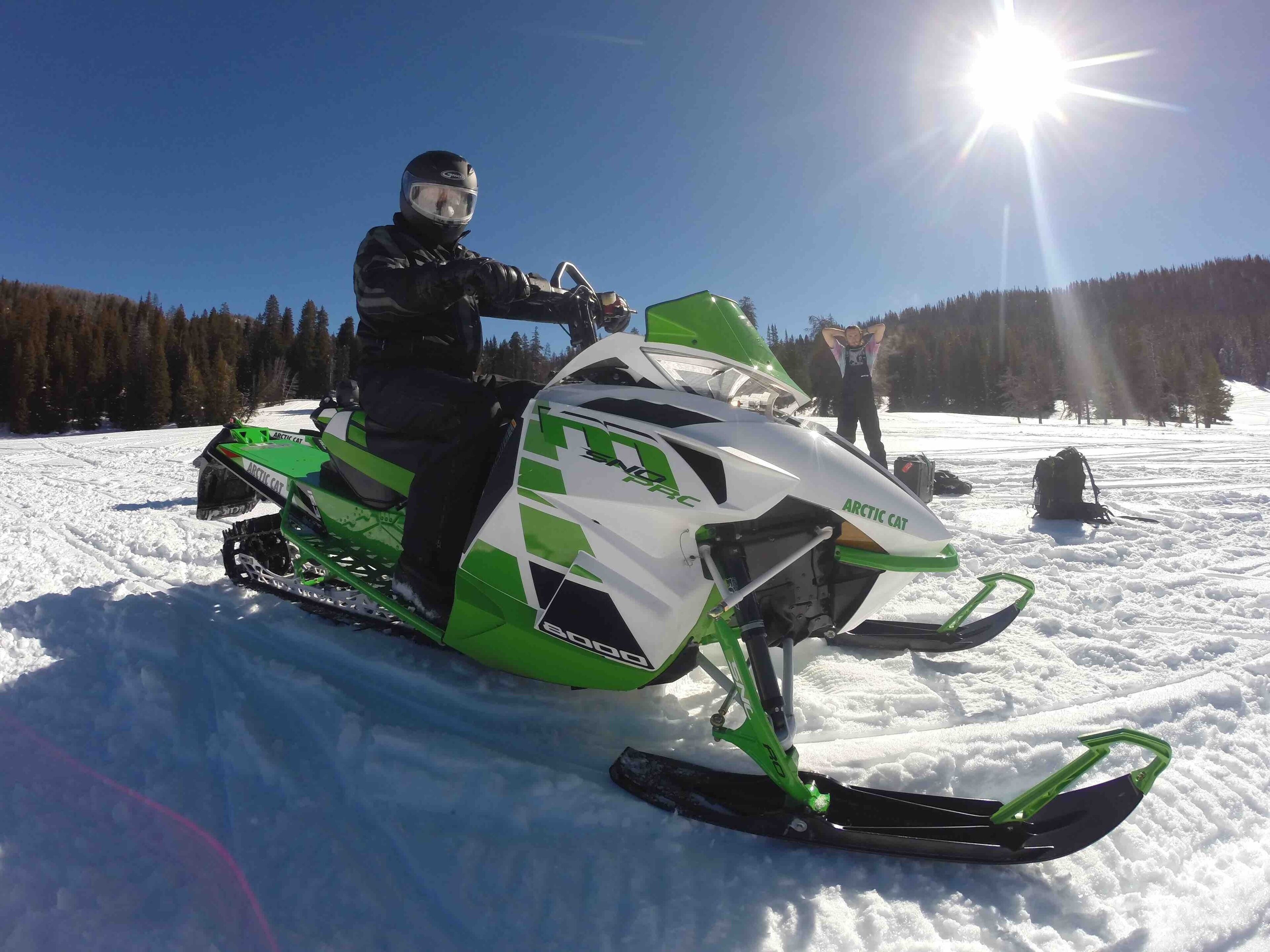 A snowmobile with a green and white color scheme, a person riding it, and a snowy landscape with trees in the background.