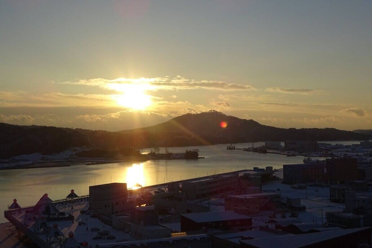 Paisagem costeira ao pôr do sol, com um corpo d'água, edifícios e uma colina ao fundo.