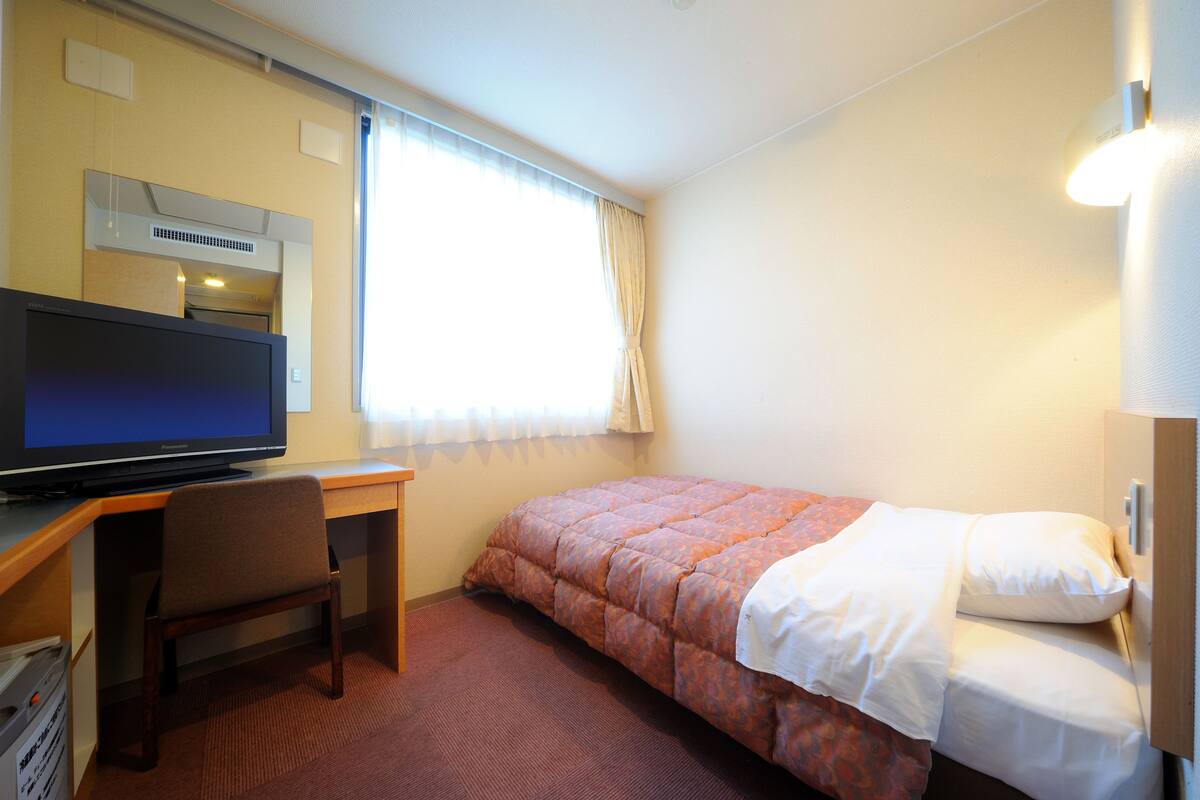 Quarto de hotel com cama, escrivaninha, televisão e janela com cortinas.