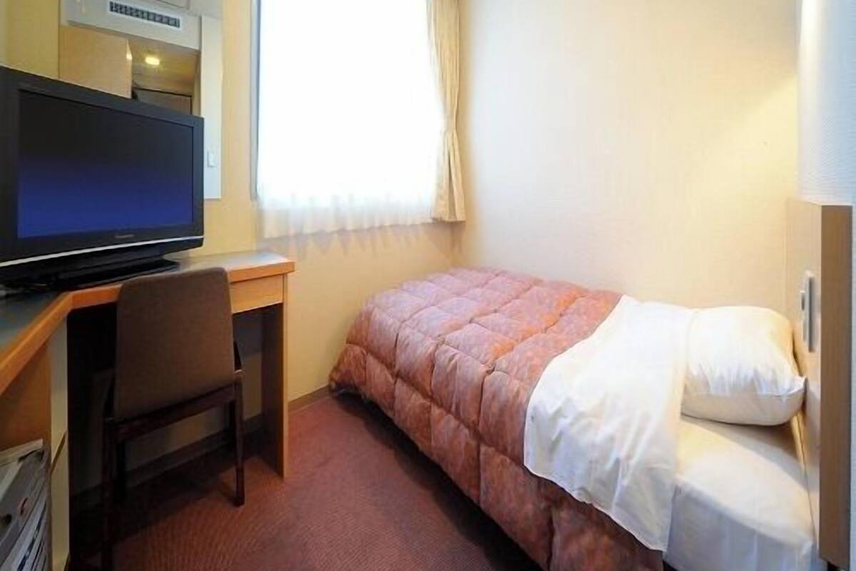 Quarto de hotel com cama, escrivaninha com televisão, cadeira e janela com cortinas.