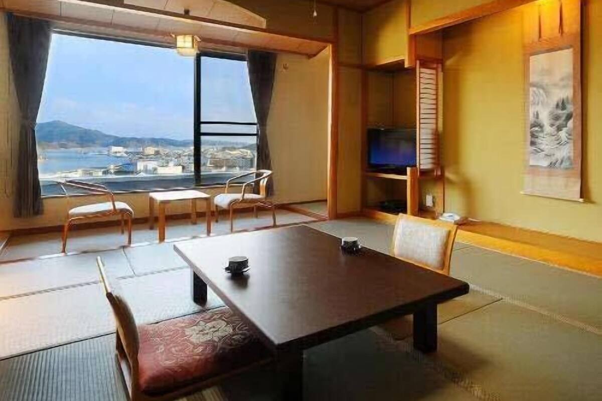 Quarto com uma mesa de madeira, cadeiras, televisão e vista para a cidade através de grandes janelas.