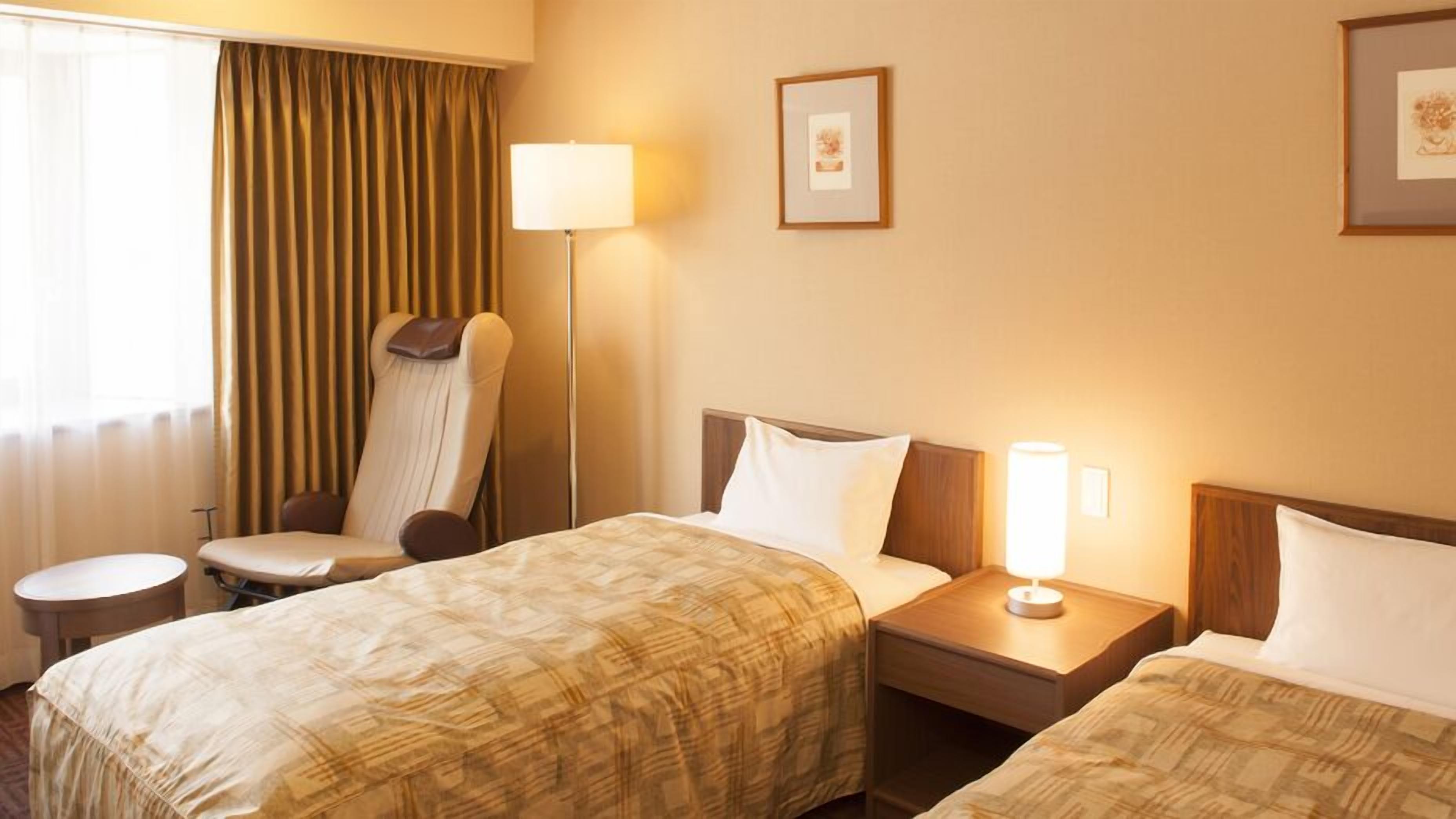 A hotel room with two beds, a chair, a bedside table, a lamp, and a window with curtains.