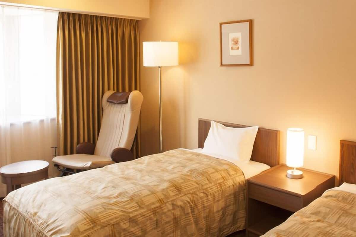 A hotel room with two beds, a chair, a bedside table, a lamp, and a window with curtains.