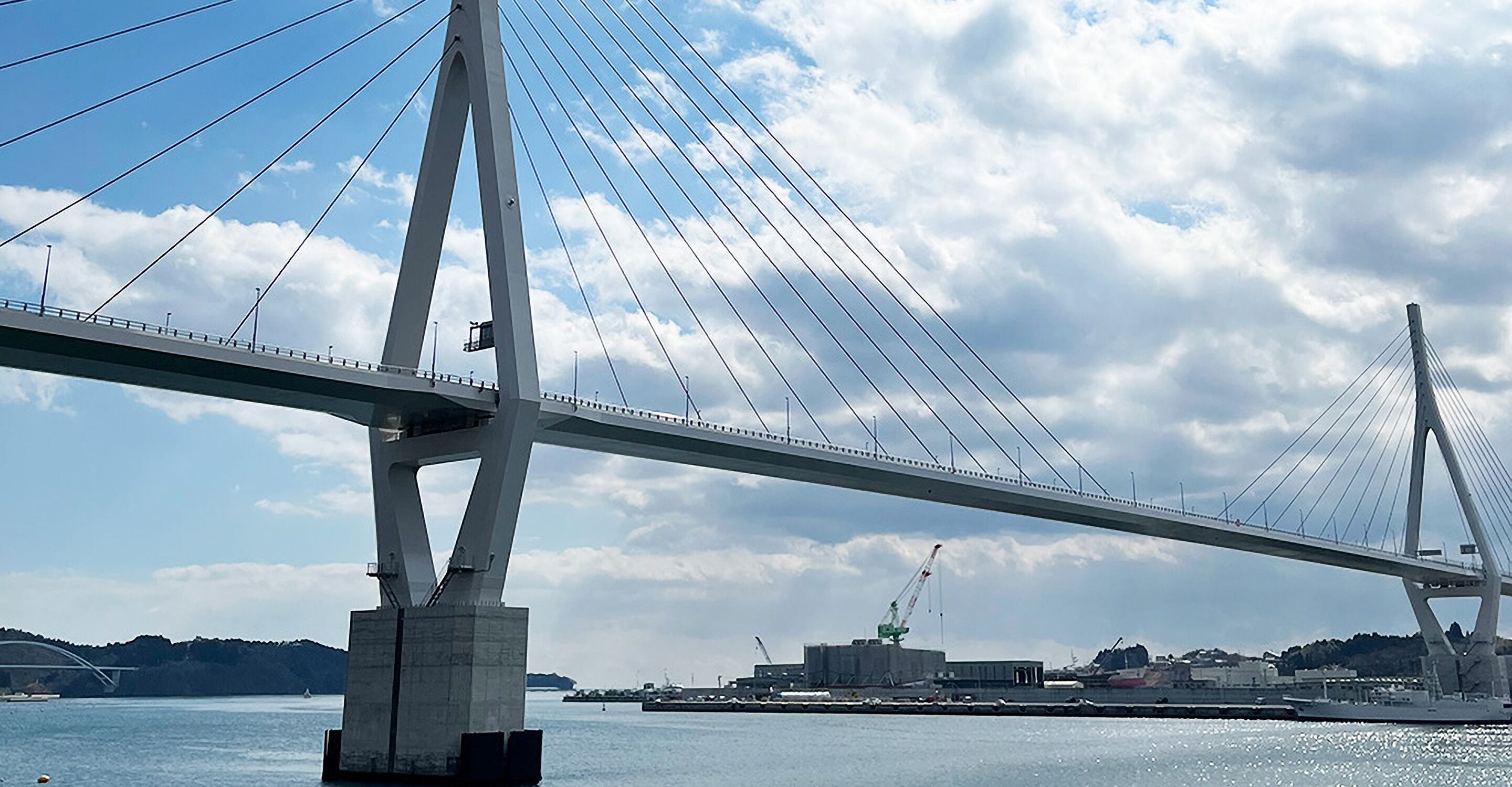 A large suspension bridge with cables and towers, spanning over a body of water with industrial buildings in the background.