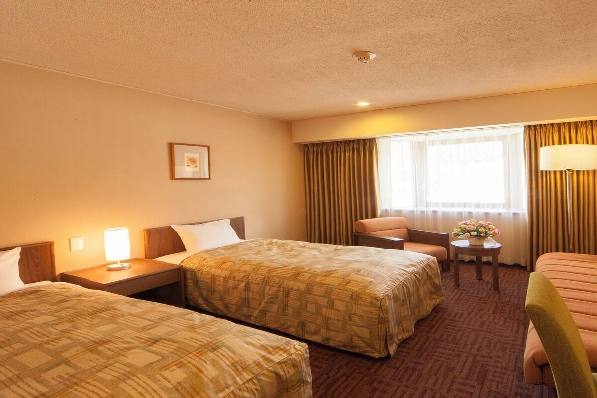 Habitación de hotel con dos camas, un sofá, una silla, una mesita con un florero y una lámpara.