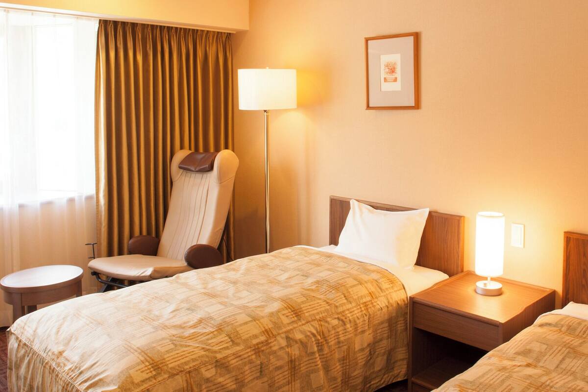 Una habitación de hotel con dos camas, una silla, una mesita de noche, una lámpara y cortinas.