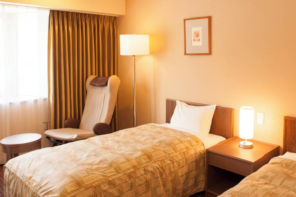 A hotel room with two beds, a chair, a bedside table, a lamp, and curtains.