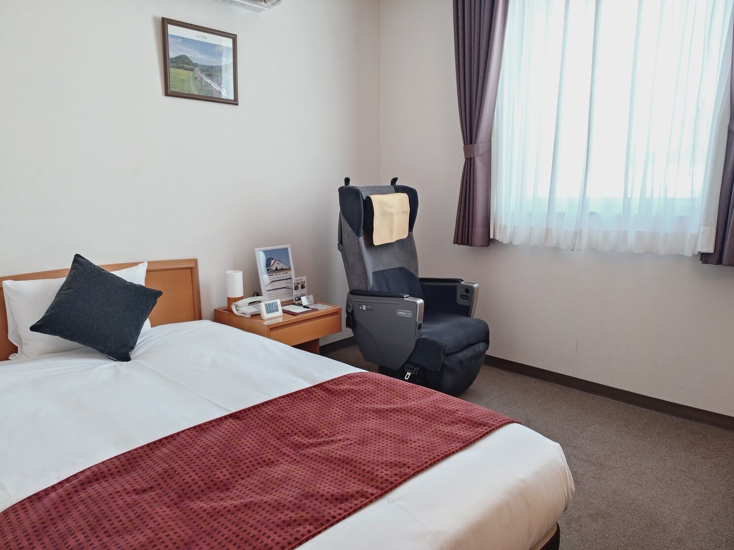A hotel room with a bed, a desk with a phone and a lamp, a chair, a window with curtains, and a framed picture on the wall.