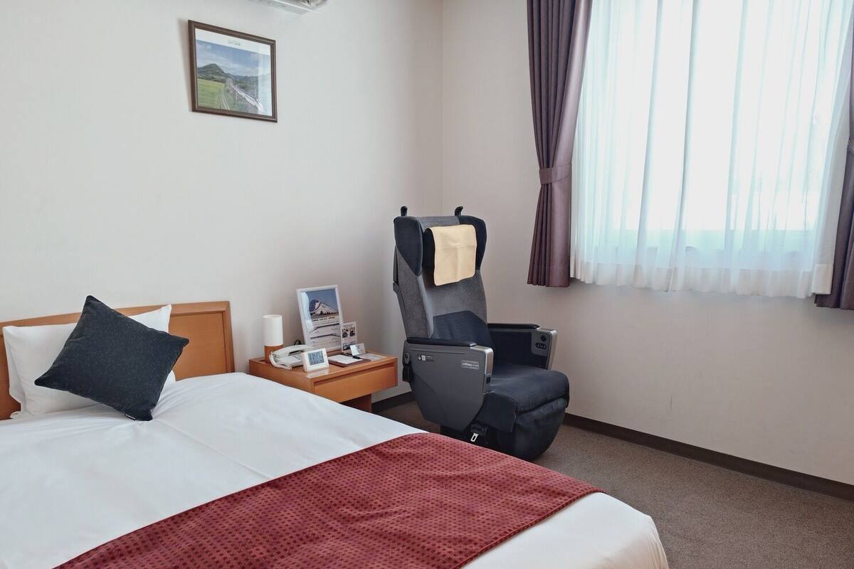 Ein Hotelzimmer mit Bett, Schreibtisch mit Telefon und Lampe, Stuhl, Fenster mit Vorhängen und einem gerahmten Bild an der Wand.