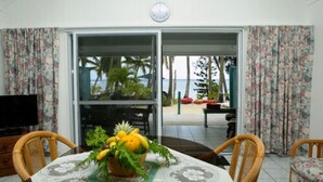 A dining area with a table set for two, a television on a cabinet, and a view of a beach through a sliding glass door. - Sunhaven Beach Bungalows (Rarotonga)