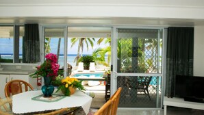 A dining area with a table set for two, a TV on a stand, and a view of a pool and palm trees through a glass door. - Sunhaven Beach Bungalows (Rarotonga)