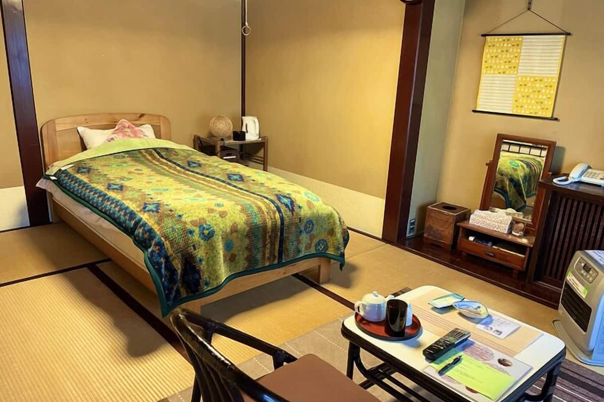 A traditional Japanese-style room with tatami flooring, a low bed, a small table with a cup and saucer, a chair, and a bedside table with a lamp and a decorative item.