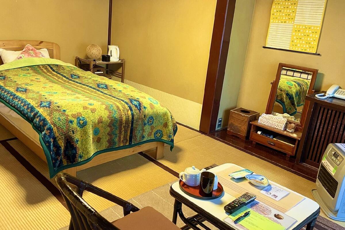 A traditional Japanese-style room with tatami flooring, a low bed with a green and blue patterned bedspread, a wooden nightstand with a lamp, a small table with a teapot, a mirror, and a telephone.