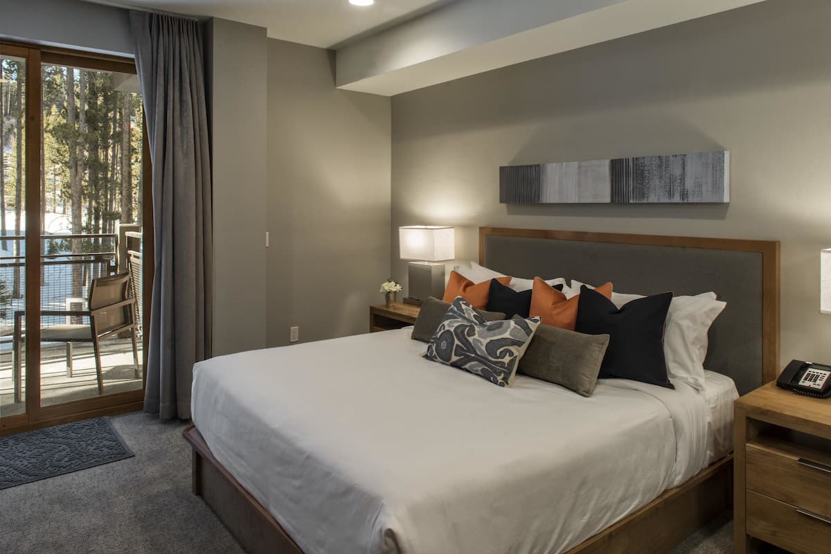 Ein modernes Hotelzimmer mit einem großen Bett, einem Nachttisch mit Lampe, einem Holz-Kopfteil und Blick ins Freie durch eine Schiebetür.