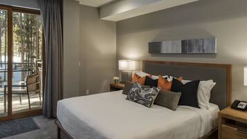 A modern hotel room with a large bed, a nightstand with a lamp, a wooden headboard, and a view of the outdoors through a sliding door.