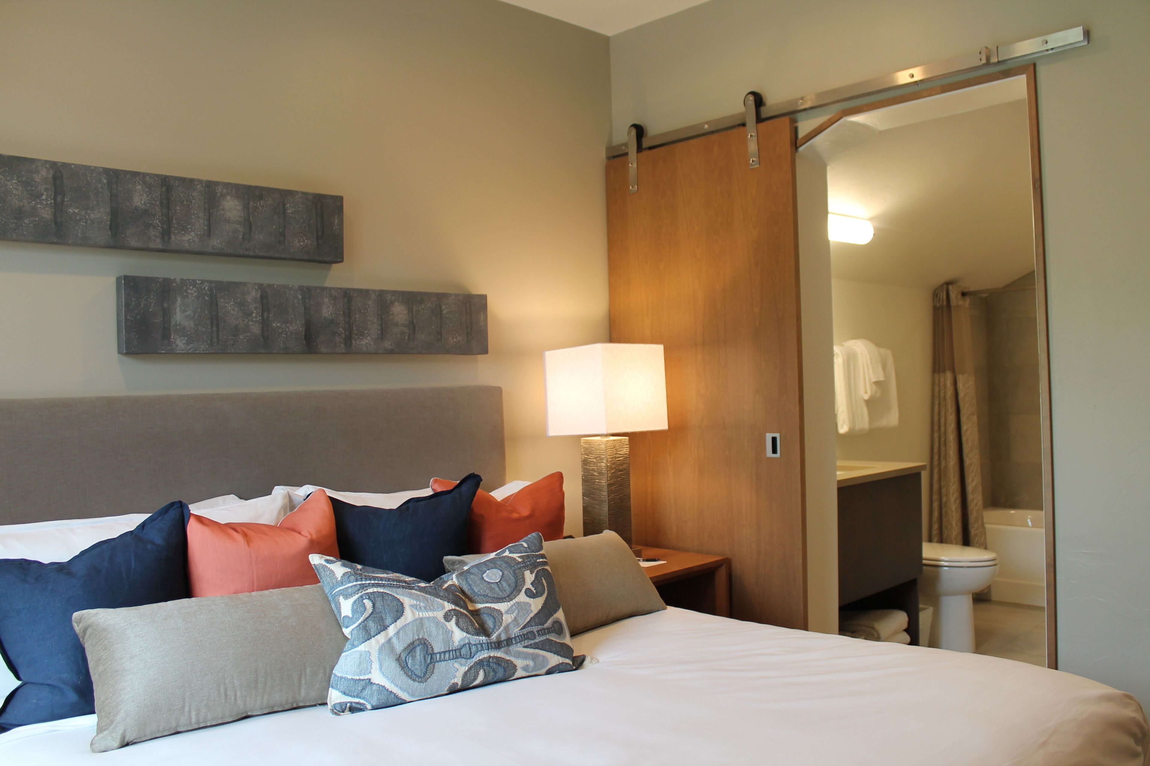 A hotel room with a bed, pillows, a nightstand, a lamp, a sliding door to a bathroom, and a mirror.